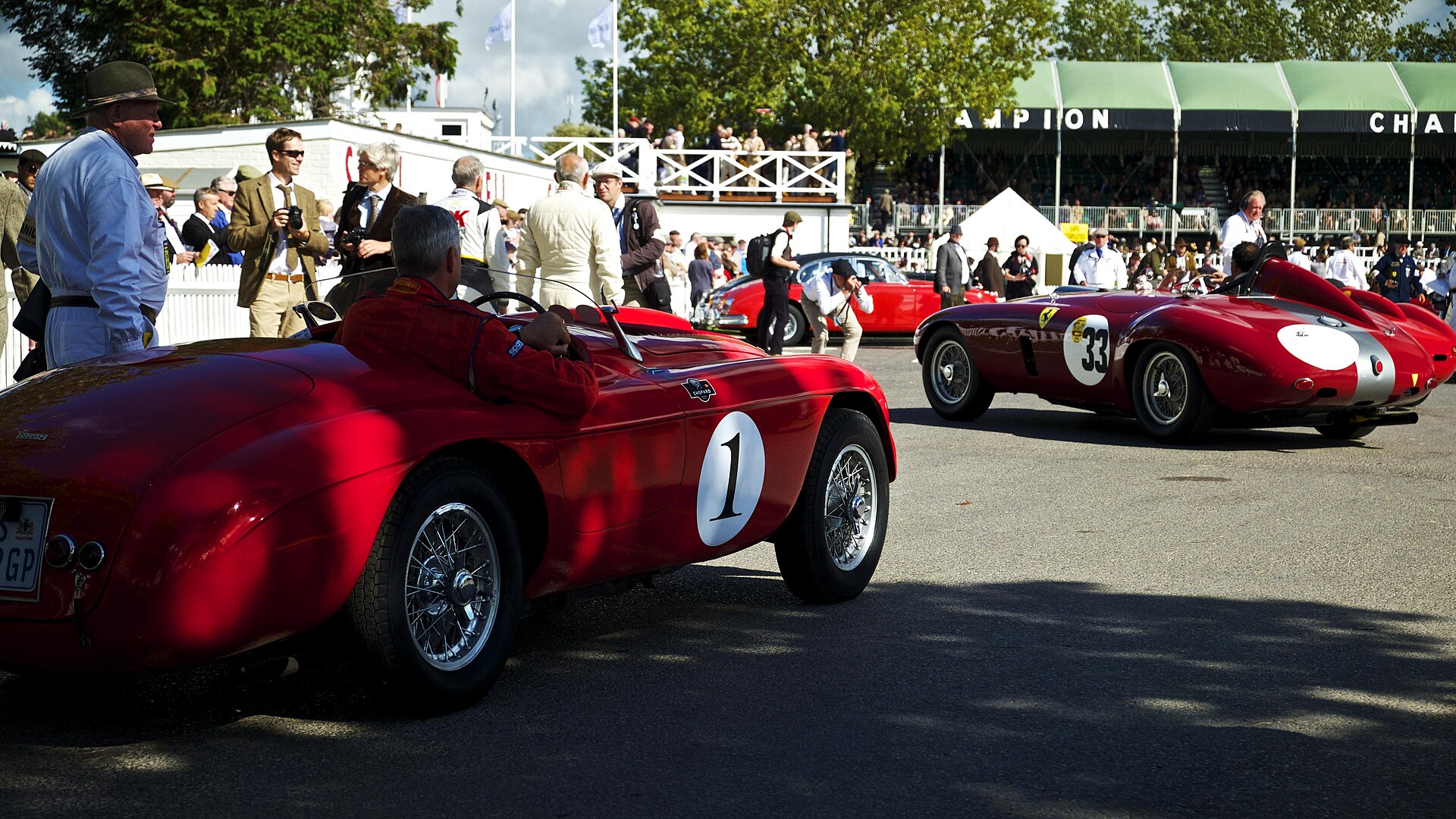 Two scarlet 1950s Ferrari sports racers wearing numbers 1 and 33 roll through the Goodwood Revival paddock past drivers and spectators in period dress.